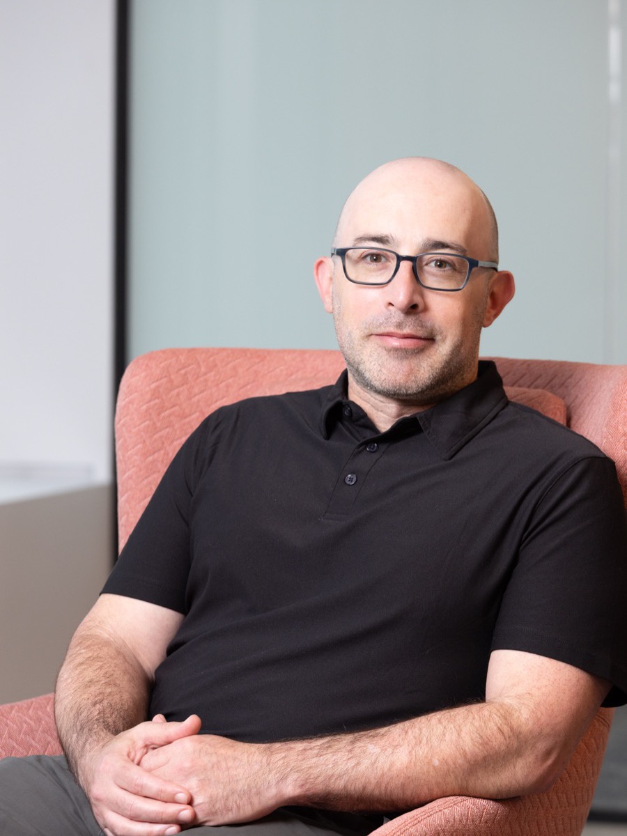 Portrait of a man with glasses in a black polo shirt, seated in a coral armchair