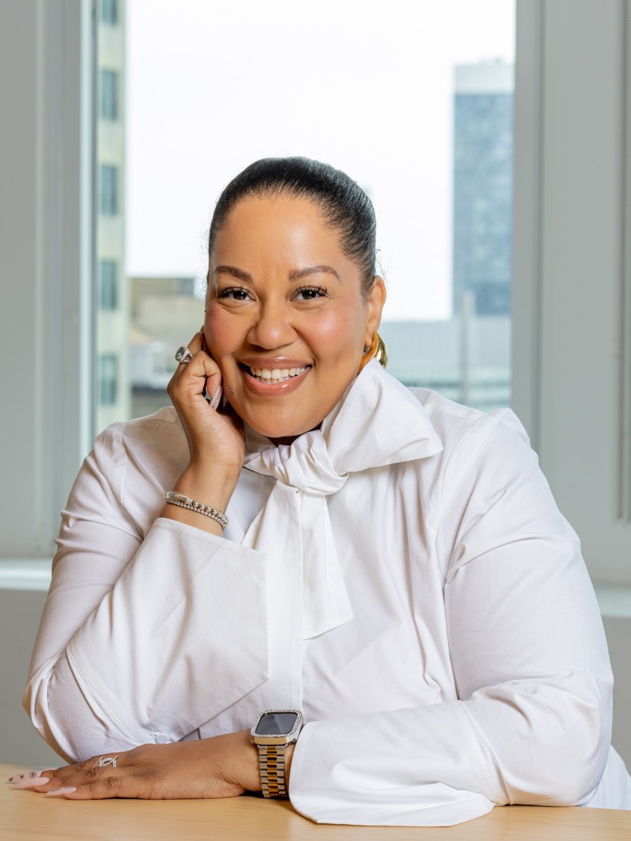 Portrait of a woman in a white pussy-bow blouse and gold hoop earrings, chin resting on her hand at a table