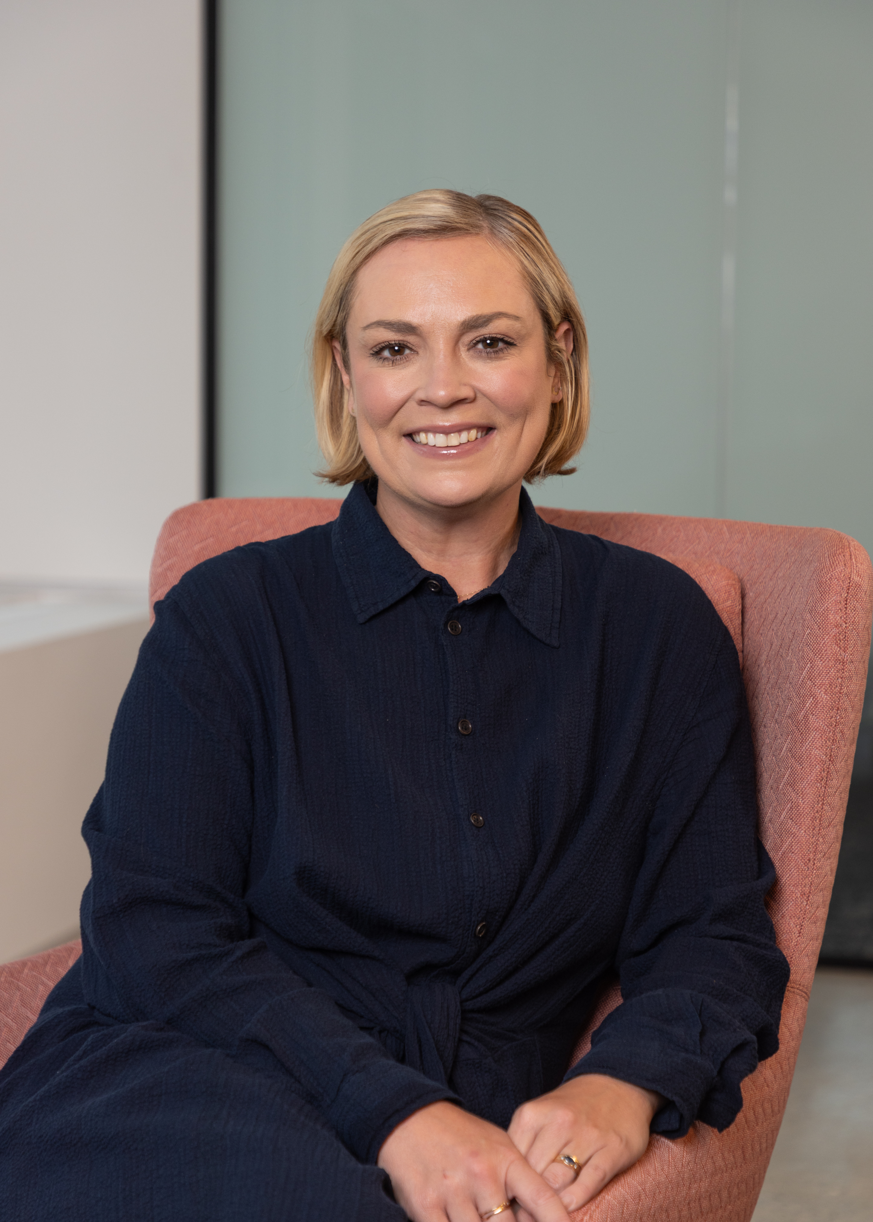 Portrait of a woman with a short blonde bob in a dark navy shirt, seated in a coral armchair by a window