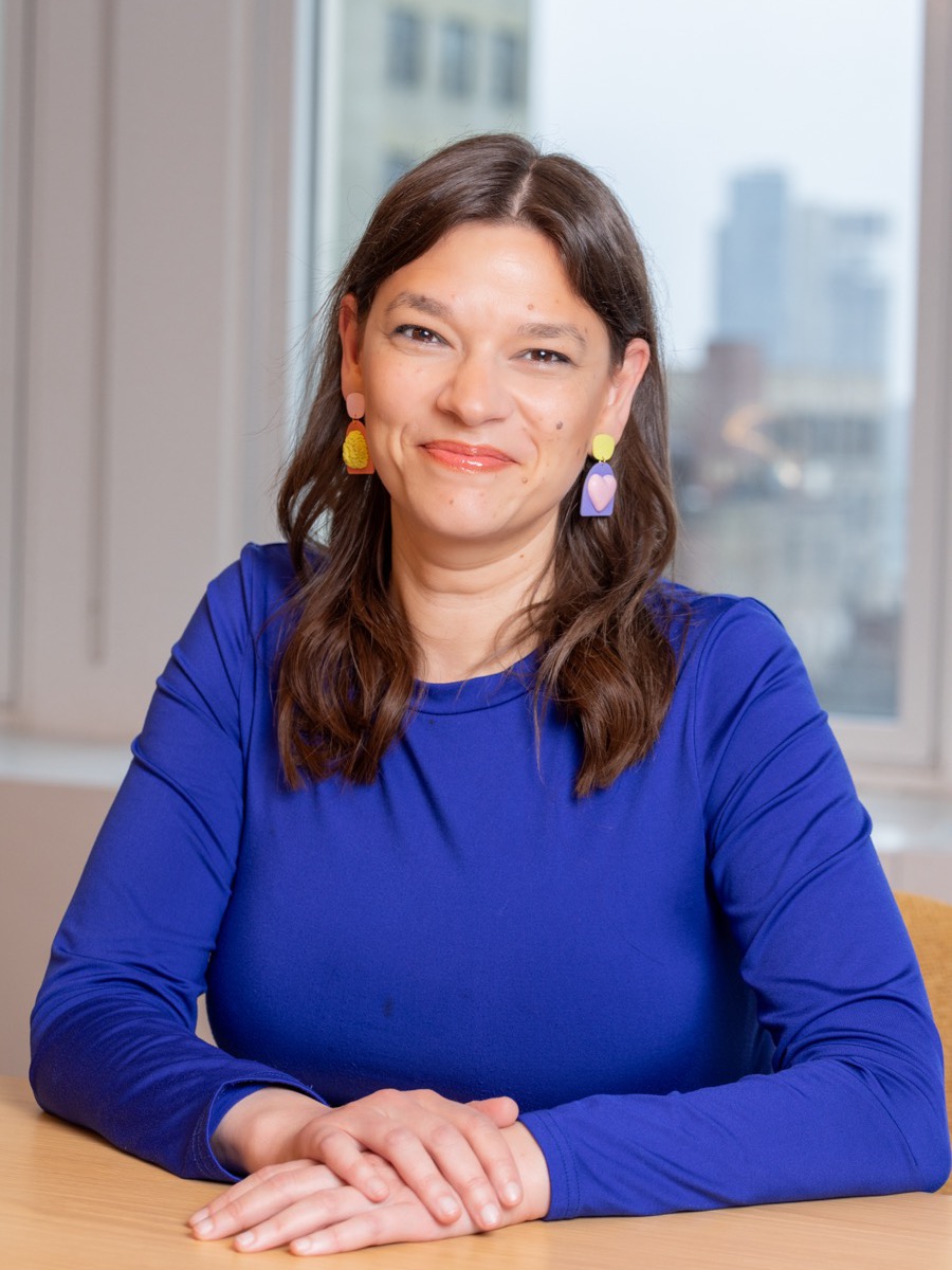 Portrait of a woman with long dark hair in a cobalt blue top and drop earrings, hands folded at a table