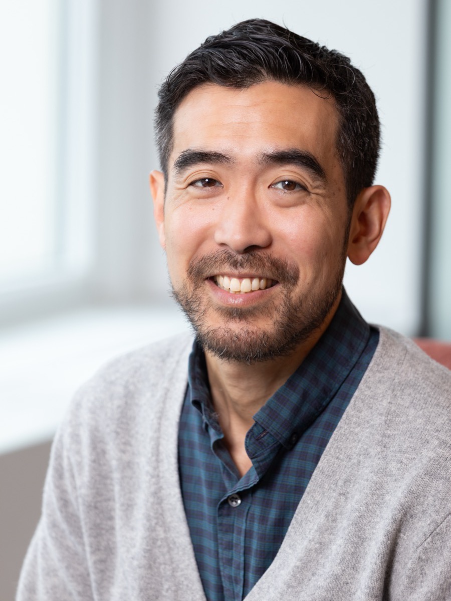 Portrait of a man in a grey cardigan over a navy checked shirt, a window behind him