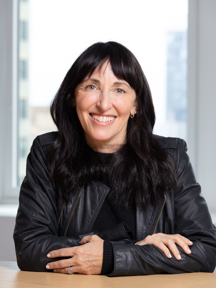 Portrait of a woman with long dark hair in a black leather jacket, arms folded at a table by a window