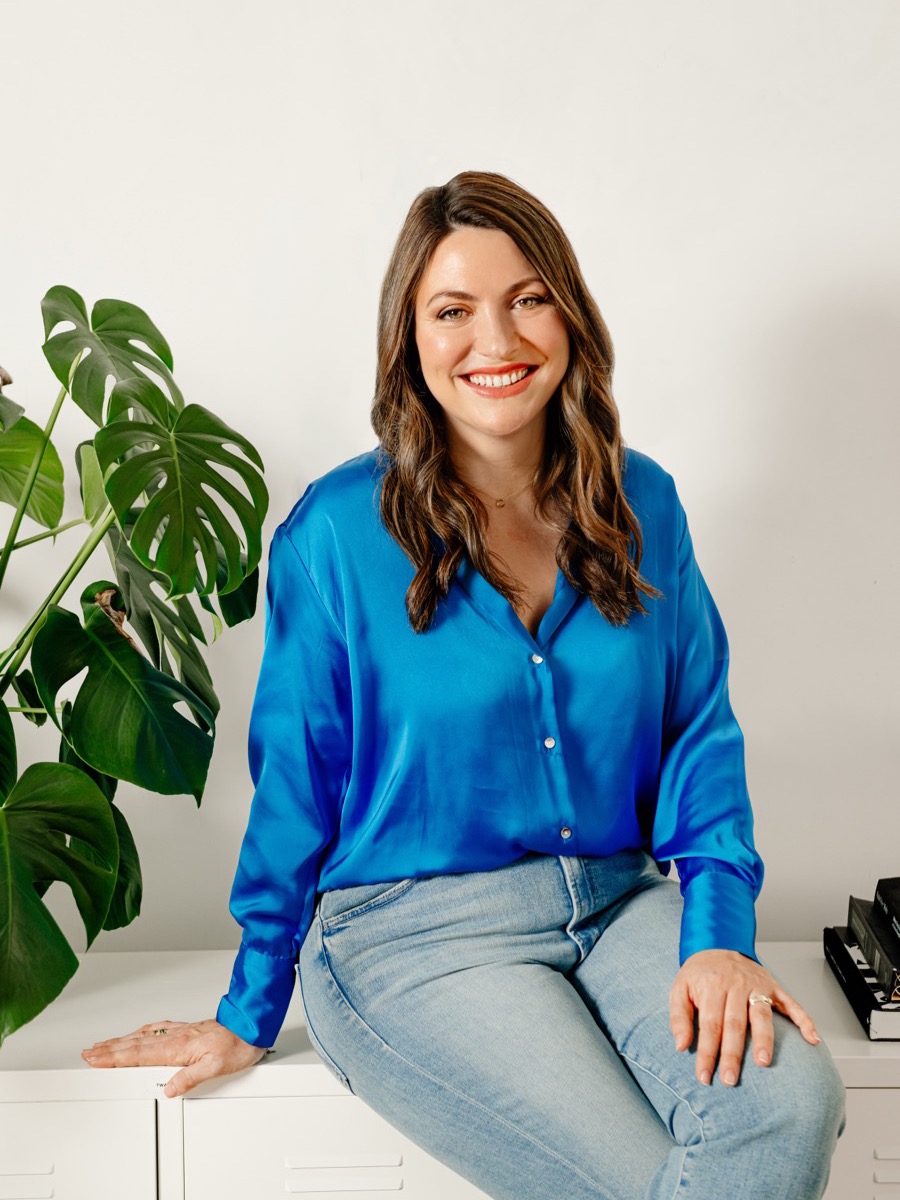 Portrait of a woman in a bright blue satin shirt and jeans, seated on a white cabinet beside a large houseplant