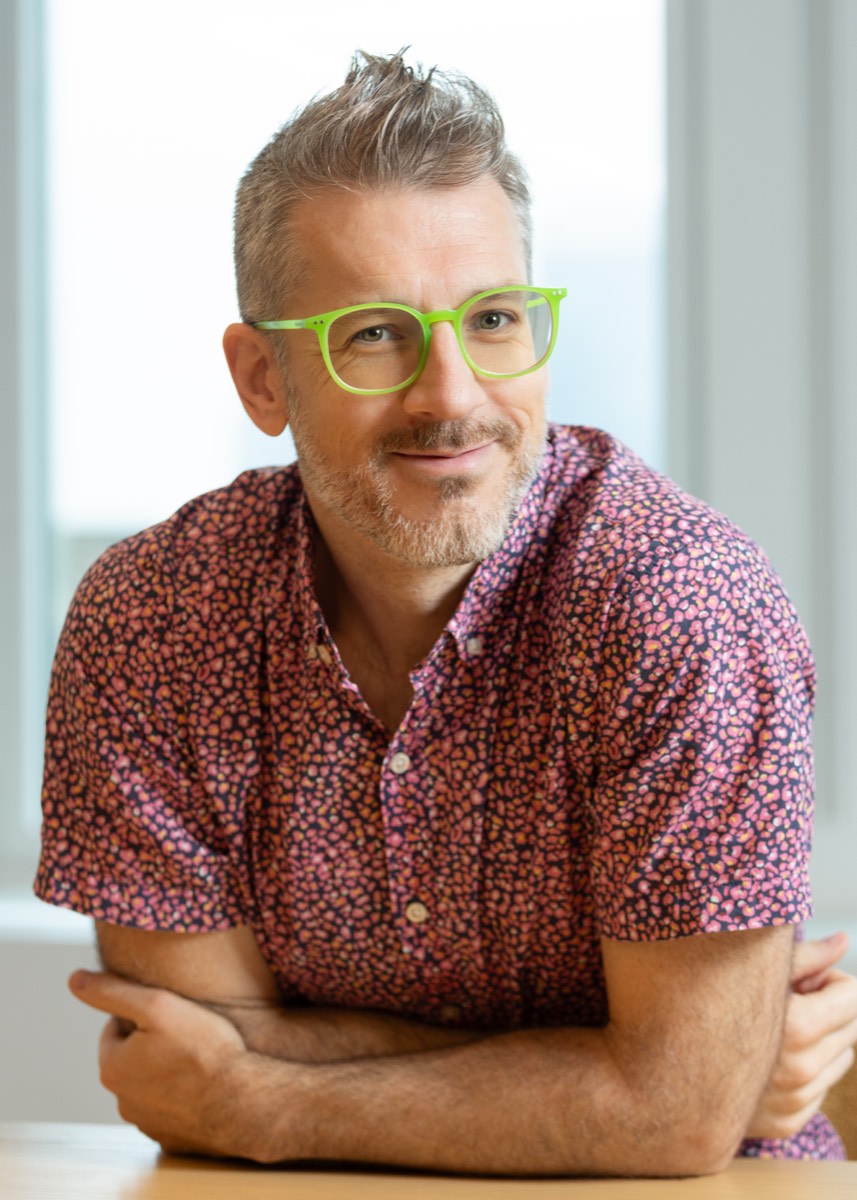 Portrait of a man with green glasses and a pink patterned shirt, arms folded at a table by a bright window