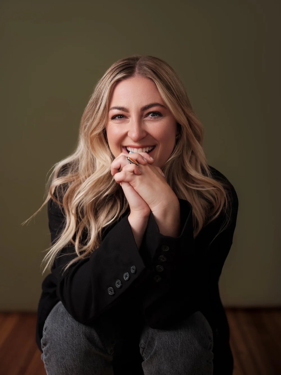 Portrait of a woman with long blonde hair in a black blazer, hands clasped by her face, against an olive backdrop