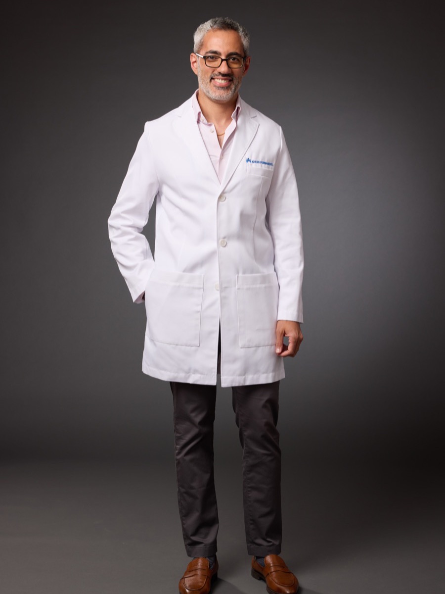 Portrait of a man in a white lab coat and glasses, standing against a dark grey backdrop
