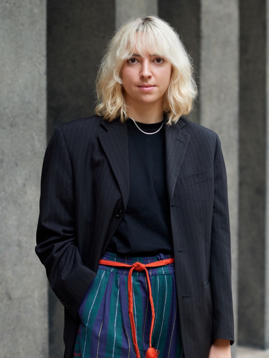 Portrait of a person with short blonde hair in an oversized black blazer and striped trousers, concrete wall behind