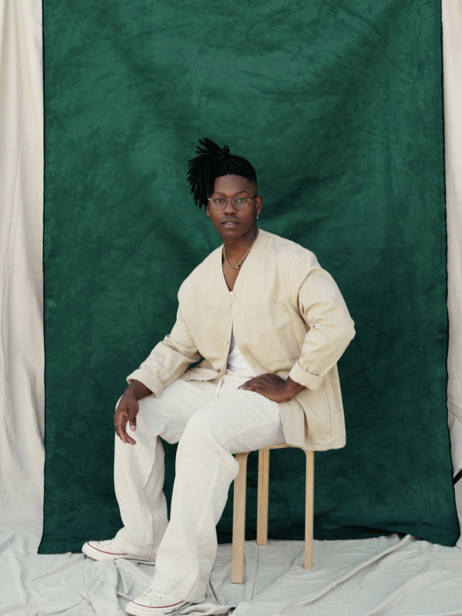 Portrait of a man in a cream suit seated on a stool, against a green fabric backdrop