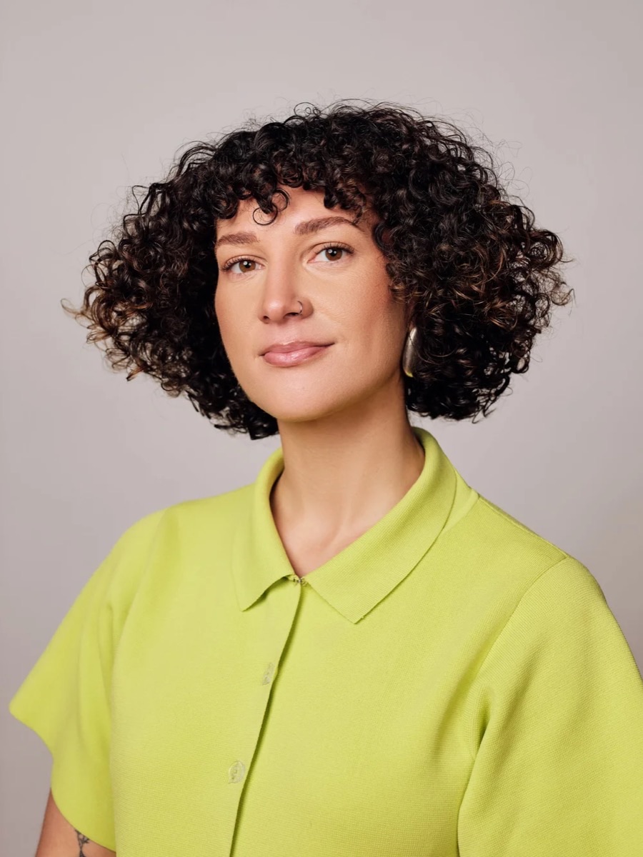 Portrait of a woman with short curly dark hair in a lime green collared shirt, against a pale grey backdrop
