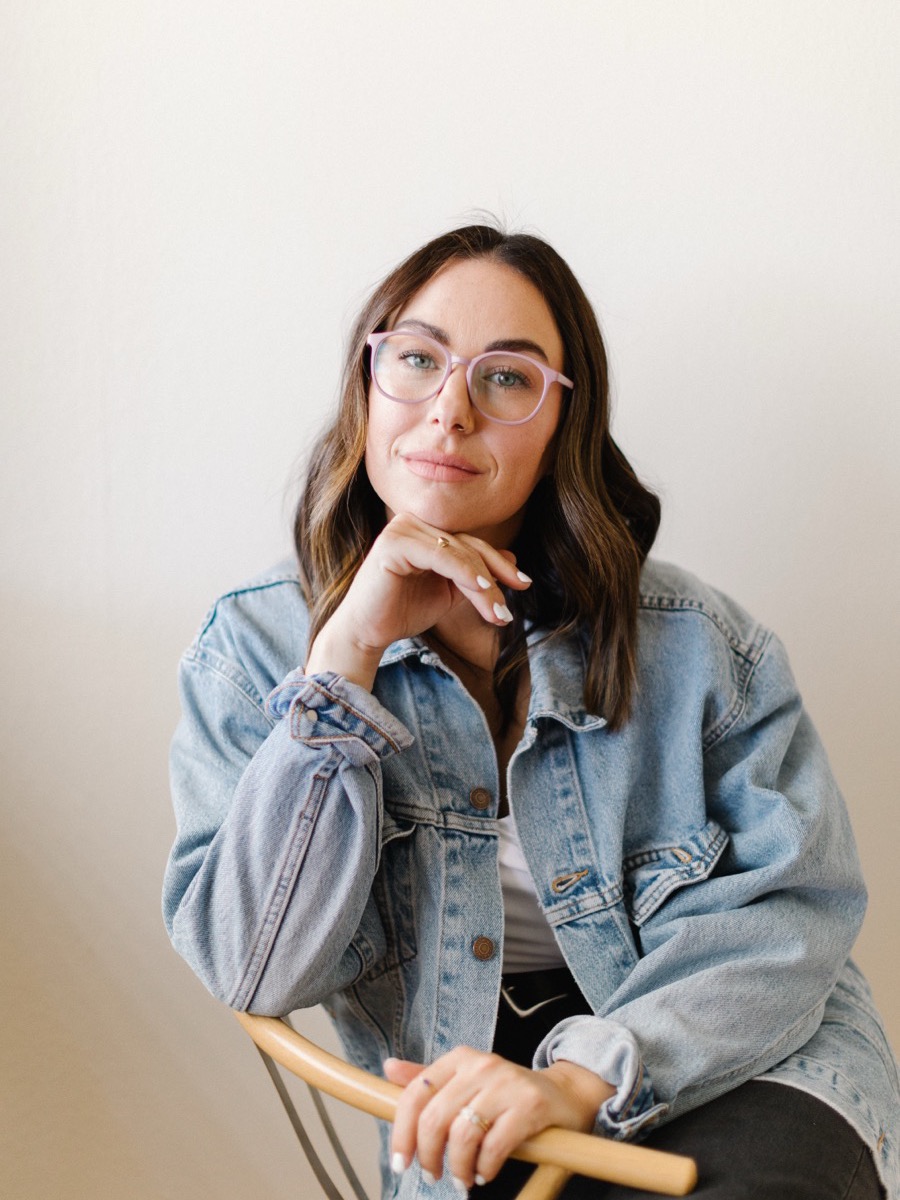 Portrait of a woman with long dark hair and glasses in a light denim jacket, chin resting on her hand, seated on a wooden chair against a pale wall
