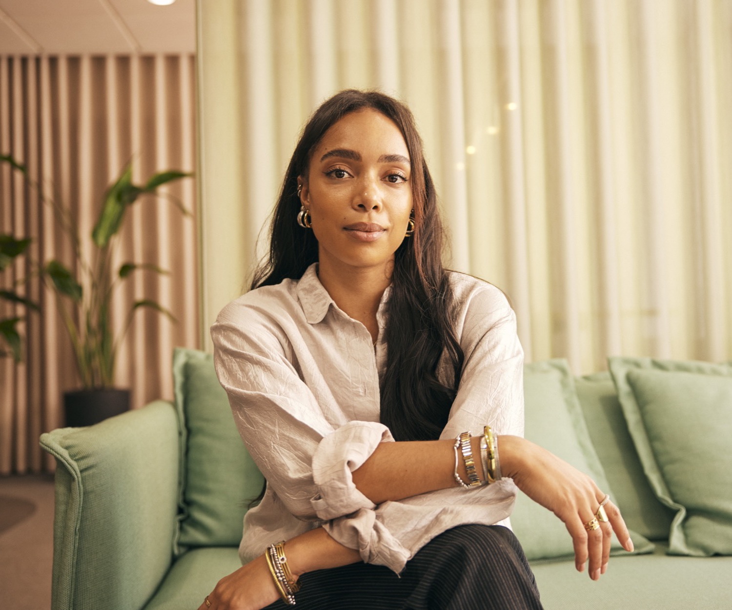 Portrait of a woman with long dark hair and gold hoop earrings in a cream linen shirt, seated on a sage green sofa beside potted plants